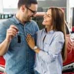 A couple is holding up keys in front of a red car at a Buy Here Pay Here Car Lot.