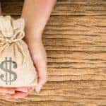 Two hands are cradling a bag of money over a wooden desk.