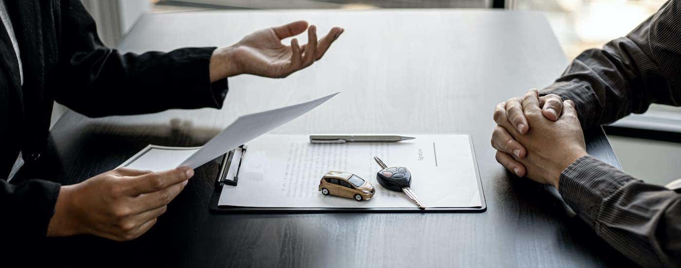 A close up shows two people at a table with a clipboard, toy car, and car keys.