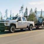 A white 2020 Chevy Silverado 2500HD is towing construction equipment on a forest highway.