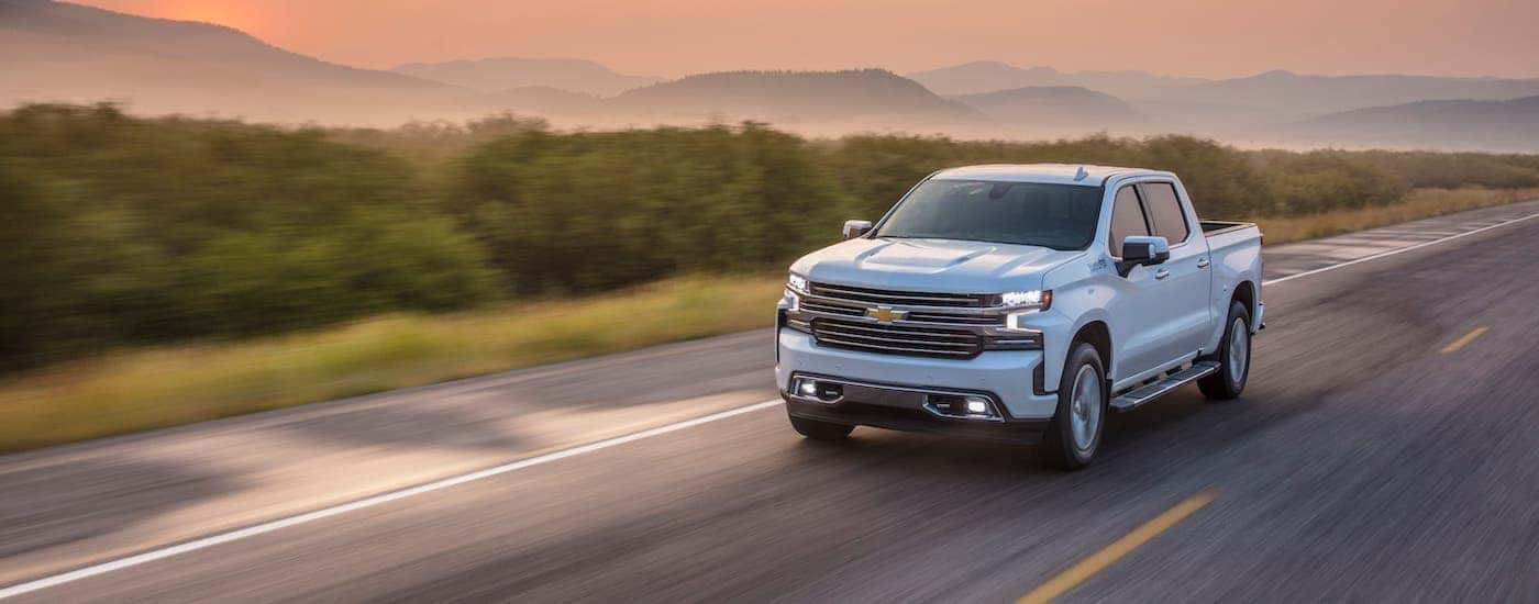 A white 2021 Chevy Silverado 1500 High Country is driving on an empty highway below a vibrant pink sunset.
