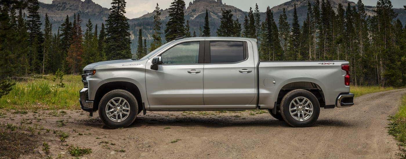 A silver 2021 Chevy Silverado 1500 LT is shown from the side while parked in front of evergreen trees and mountains.