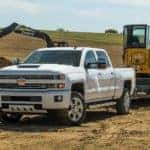 A white 2018 Chevy Silverado 2500HD is towing a front loader through a construction site.