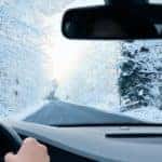 A snowy tree-lined road is shown through the windshield of a vehicle.