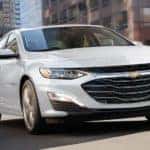 A white 2021 Chevy Malibu is shown from the front while driving down a city street.