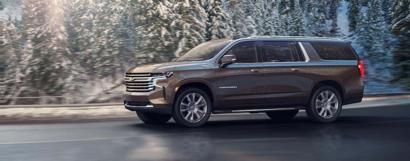 A grey 2021 Chevy Suburban High Country is shown from the side driving past snow covered trees.