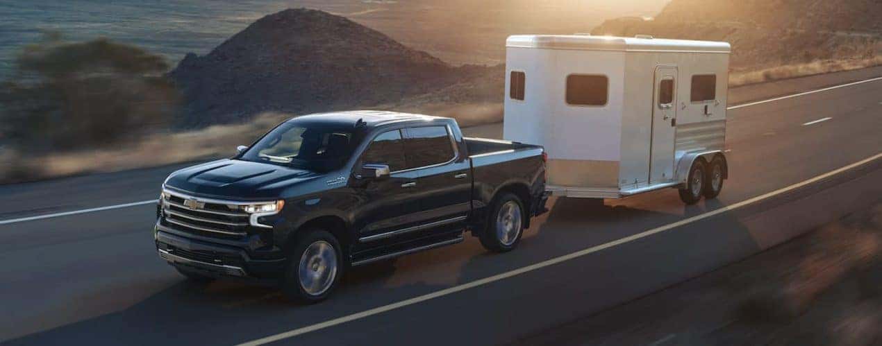 A black 2023 Chevy Silverado 1500 is shown towing a trailer on an open road.