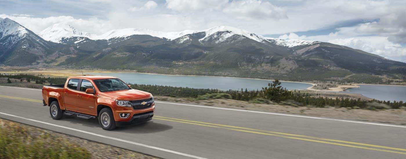 An orange 2016 Chevy Colorado Z71 is shown driving past a lake and snow covered mountains.