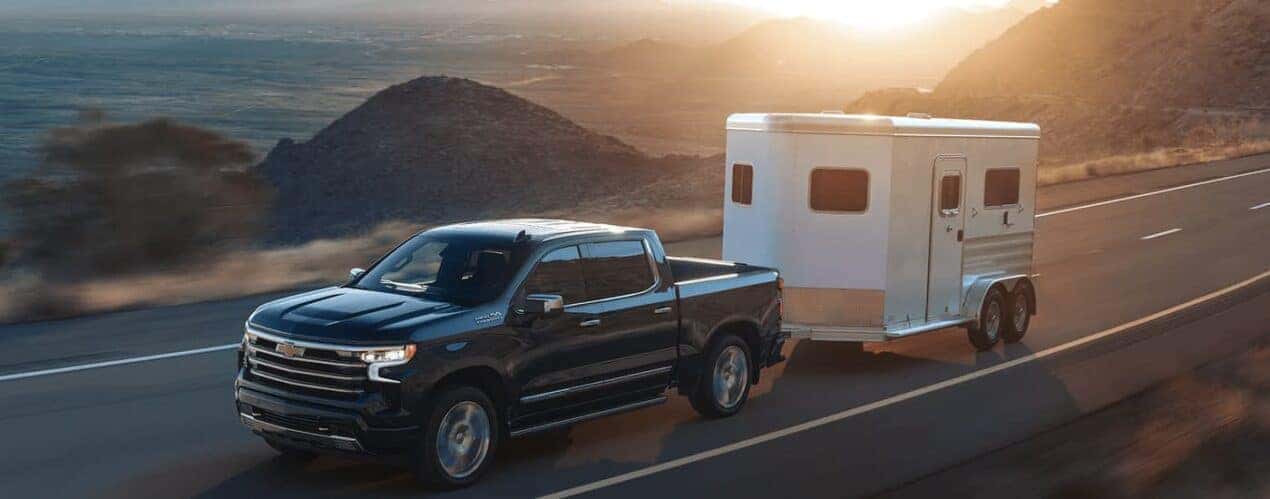 A black 2023 Chevy Silverado 1500 High Country is shown towing a white trailer on an open highway.