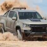 A tan 2021 Chevy Colorado ZR2 is shown from the front at an angle while driving through sand after leaving a Certified Pre-Owned Chevy dealer.
