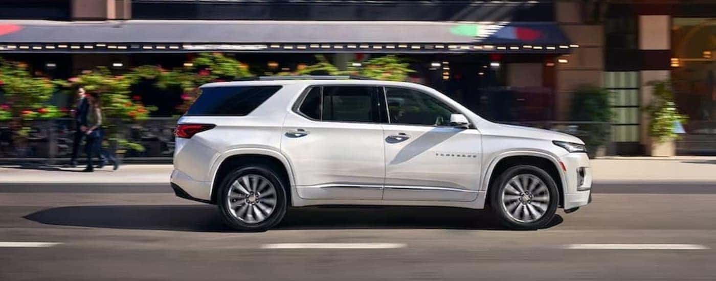 A white 2023 Chevy Traverse is shown driving on a city street after visiting a used SUV dealer in Indianapolis.