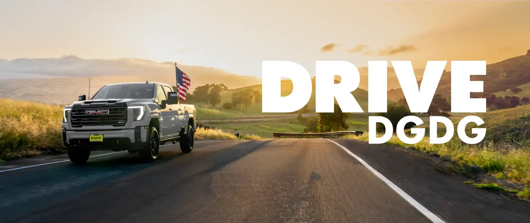 Silver GMC Sierra 1500 AT4 pickup truck flying an American flag from its bed while driving along a country road at sunset.