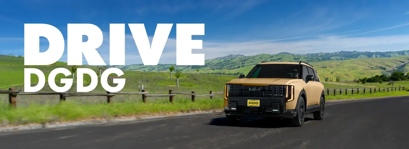 Tan Kia Telluride SUV driving along a paved road with rolling green hills and a bright blue sky in the background.