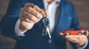 A man in a blue suit is holding out a set of car keys with one hand and has a toy car in the other hand.