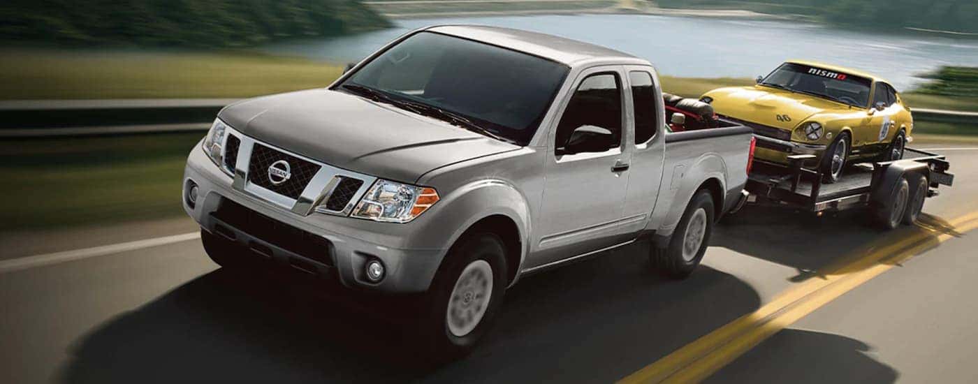 A silver 2020 Nissan Titan is towing a vintage yellow Nissan past a lake.