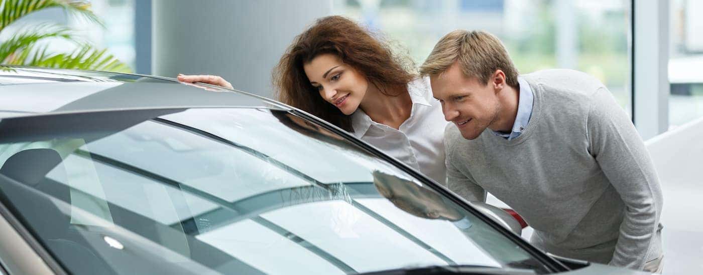 Two people are shown at a car dealership looking into the window of a car. 