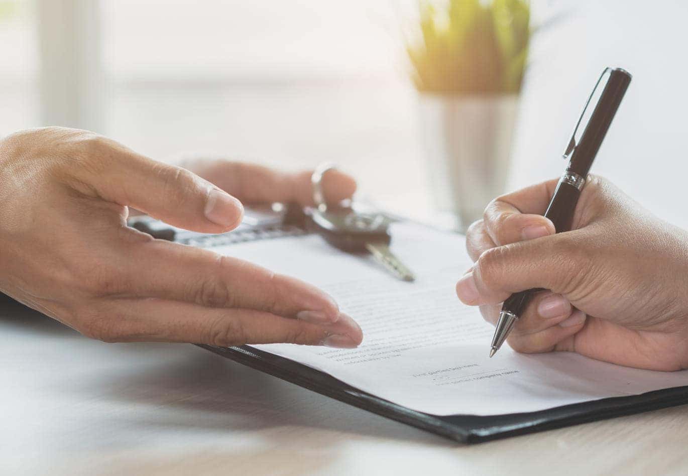 Close-up of hands exchanging car keys and signing a contract on a clipboard, symbolizing a formal agreement.