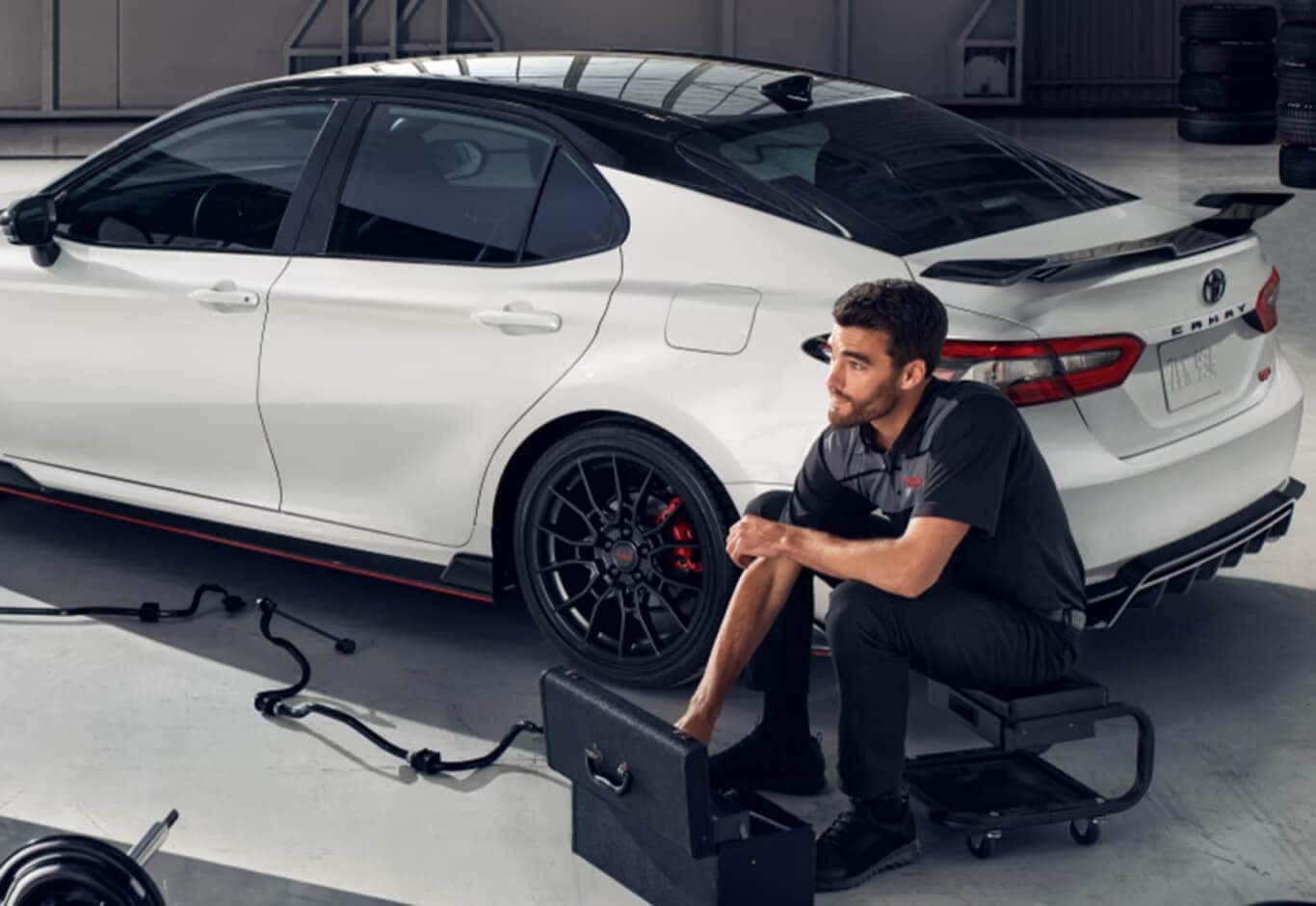 Three-quarter rear view of a Toyota Camry TRD with a mechanic sitting on a roller stool in a garage.