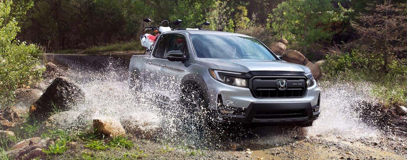 Grey 2026 Honda Ridgeline driving off-road over a shallow river.