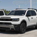 White 2025 Honda Passport TrailSport SUV in a dealership lot, showing rugged black cladding, roof rails, off-road tires, and a revised front grille.