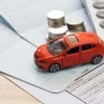 An orange toy car is shown near paperwork, coins, and a calculator.