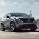 A silver 2024 Nissan ARIYA Platinum is shown driving near a city.