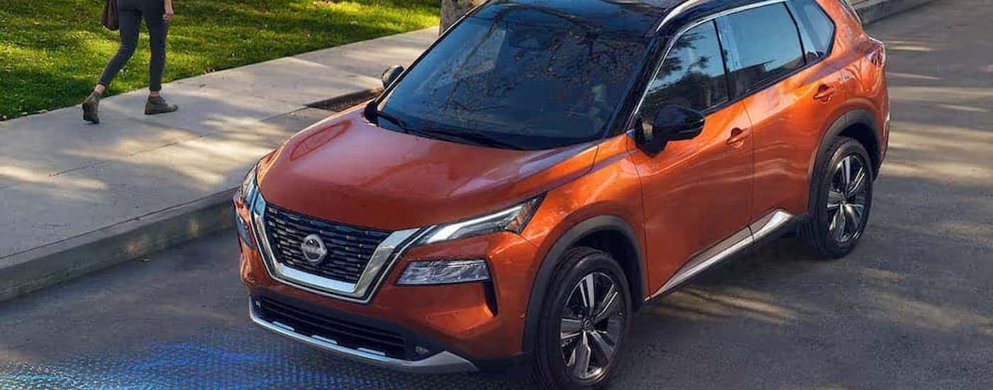 Orange 2022 Nissan Rogue driving next to a sidewalk.