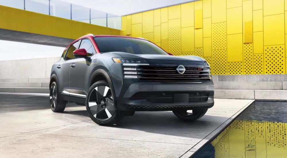 A gray 2026 Nissan Kicks SR with a red roof and side mirrors parked by a yellow building.