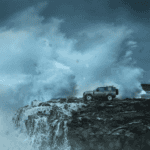 Land Rover Defender parked on rocky sea bank with wave crashing towards it
