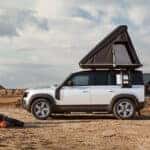A silver Defender 110 in a desert with a tent on top being used as a campsite for a family.