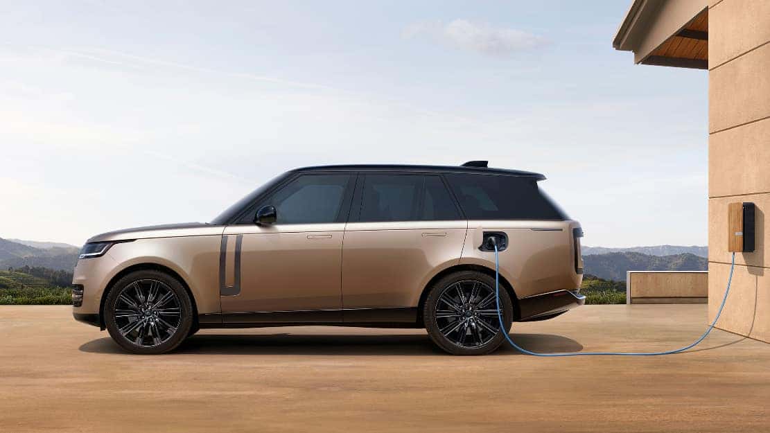 A tan Range Rover electric model in profile, plugged into a wall and charging.
