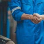 Two mechanics in blue jumpsuits shaking hands while working on a car.