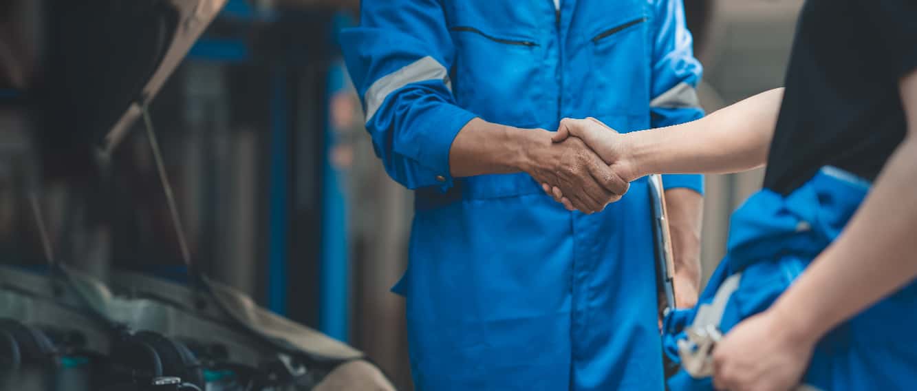 Two mechanics in blue jumpsuits shaking hands while working on a car.