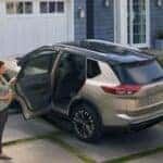 A woman stands beside the open passenger door of a 2024 Nissan Rogue parked in the driveway of a home. 2 children are nearby