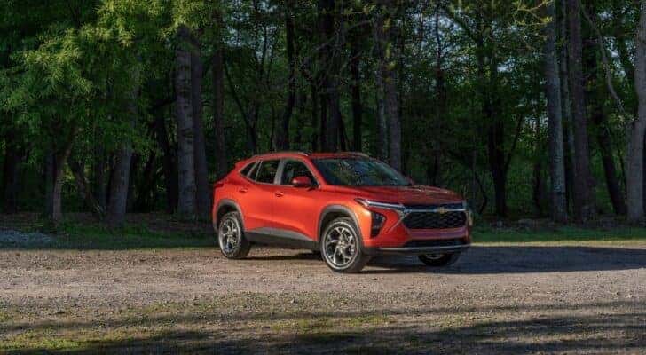 A red 2024 Chevy Trax for sale is shown on a tree-lined lot.