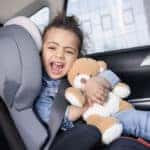 Little girl buckled into child's seat in back of car smiling and holding teddy beat