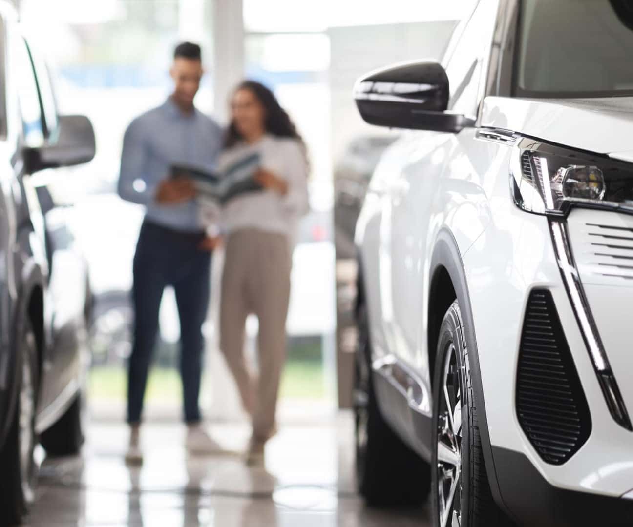 Close-up of a white SUV on display inside a dealership showroom with two shoppers in the background.