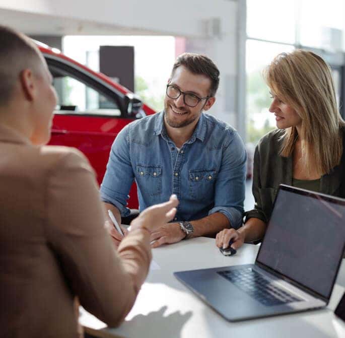 A sales associate assists a smiling couple at a desk in a bright, clean dealership.