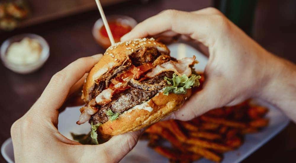 Hands are holding a gourmet burger over a plate with fries.