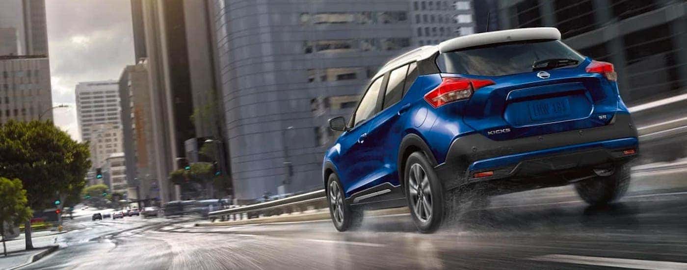 A blue 2020 Nissan Kicks is driving on a wet city street and shown from the rear.