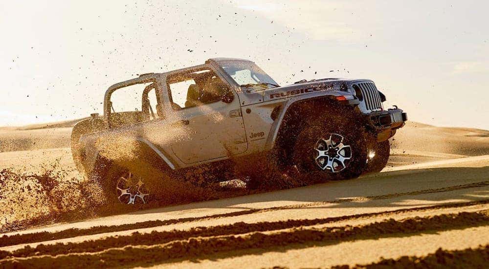 A silver 2020 Jeep Wrangler is kicking up sand while driving on a dune.