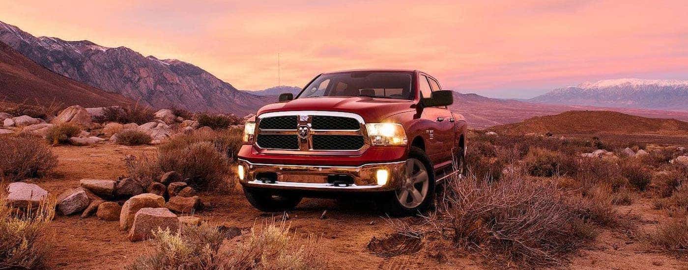 One of the older Ram trucks, a red 2019 Ram 1500 Classic, is parked in dry grass in front of mountains and a vibrant pink sunset.