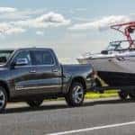A gray 2021 Ram 1500 is towing a boat on a road in front of a river.
