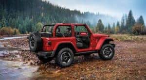A red 2019 Jeep Wrangler is parked on a rocky river bank in a forest.