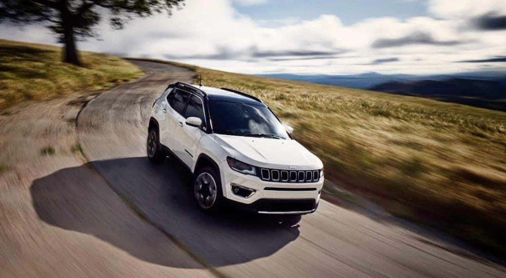 A white 2019 Jeep Compass from a used Jeep dealer is driving on a rural road past a field.
