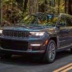 A dark grey 2021 Jeep Grand Cherokee L is shown driving down a wood-lined road.