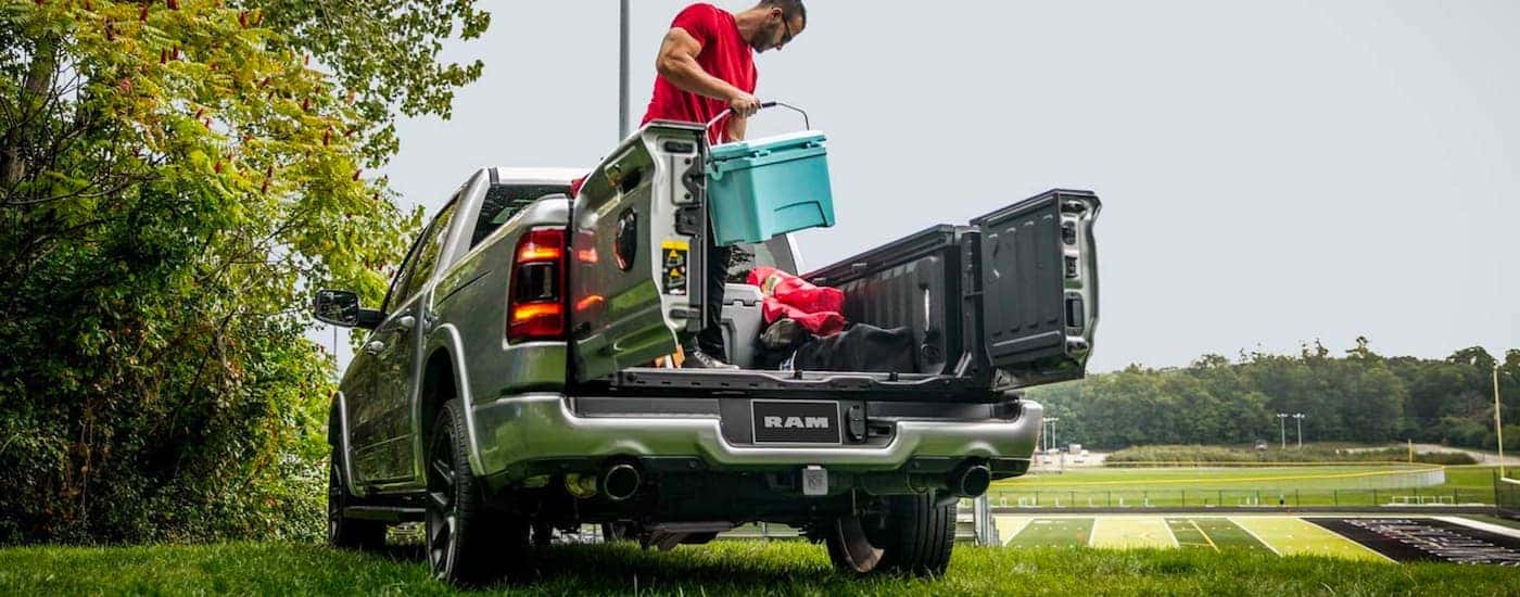 A person is shown holding a cooler with the multi function tailgate open on a 2021 Ram 1500 parked near a football field.