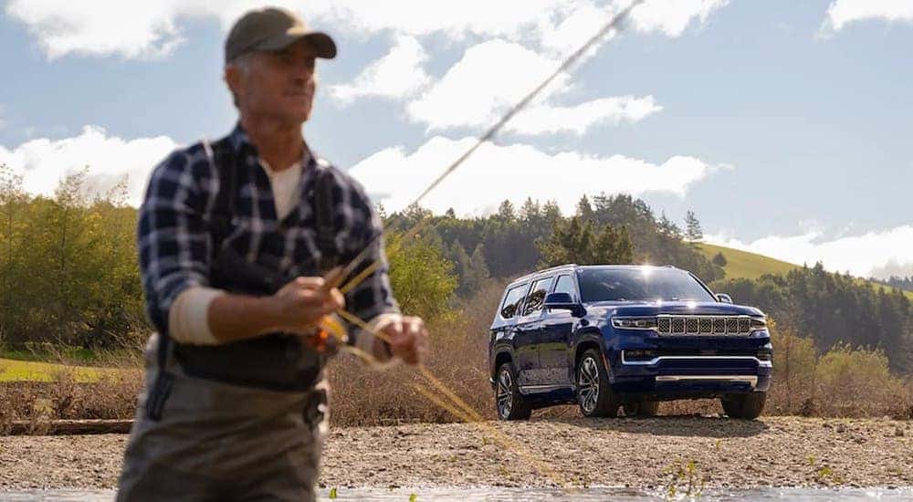 A blue 2022 Wagoneer is shown parked next to a river and a man fly fishing.