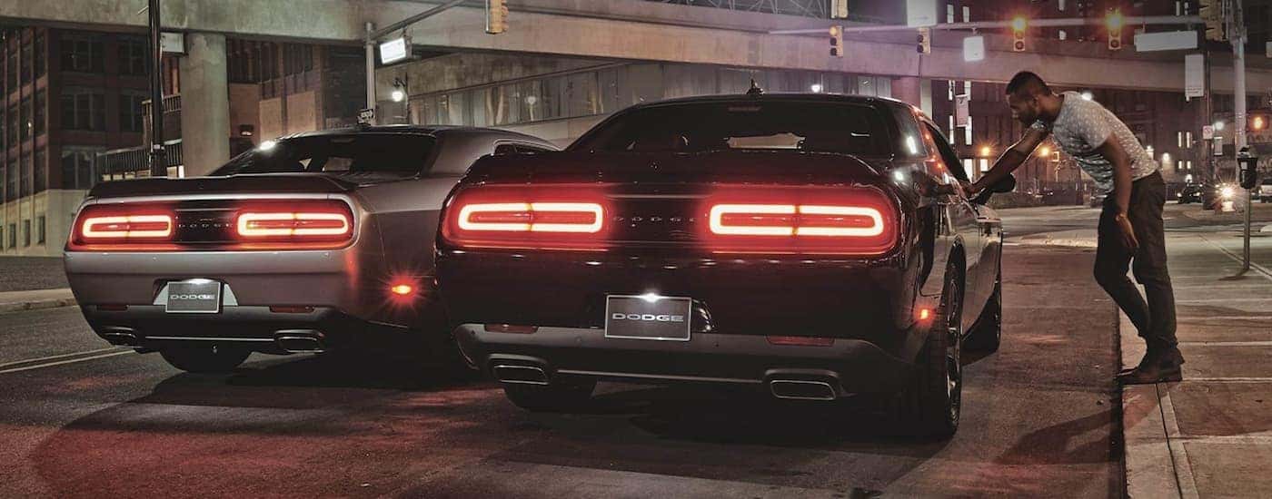 A grey and a black 2020 Dodge Challenger are shown from the rear on a city street at night.