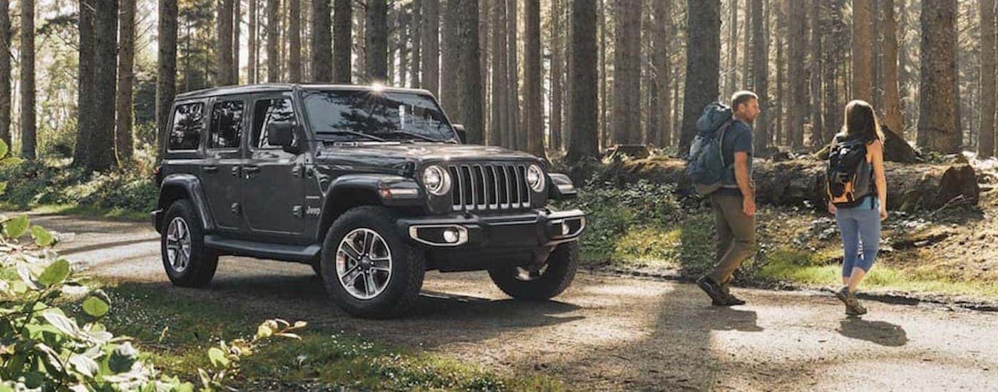 A grey 2020 Jeep Wrangler is shown parked on a dirt road as two hikers venture into the forest.